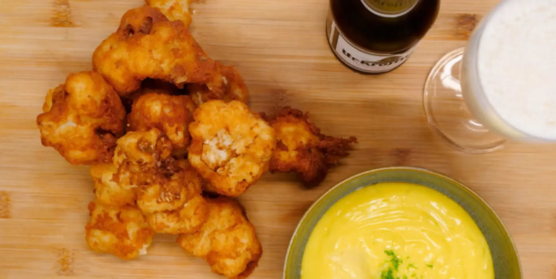 Frittierte Blumenkohlstücke liegen neben einer Schüssel gelber Soße und einem Glas Bier.