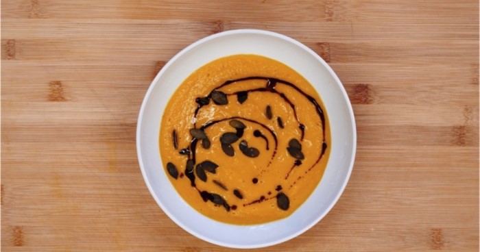 Eine Schale mit Kürbissuppe steht auf einem Holztisch, garniert mit schwarzen Kernen.