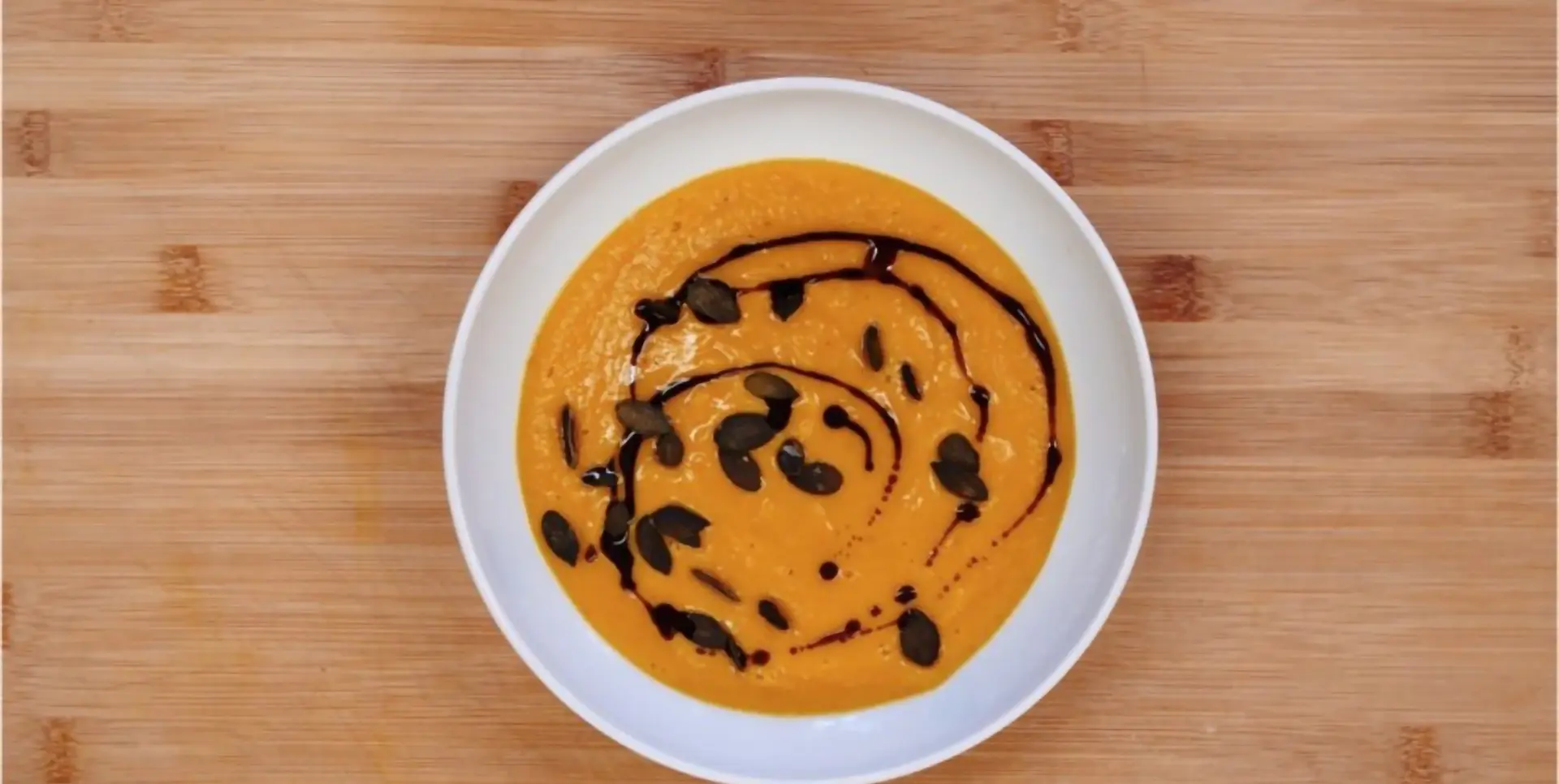 Eine Schale mit Kürbissuppe steht auf einem Holztisch, garniert mit schwarzen Kernen.