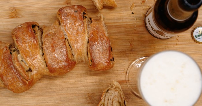 Ein geschnittenes Brot liegt neben einem vollen Bierglas und einer Flasche auf einem Holzbrett.