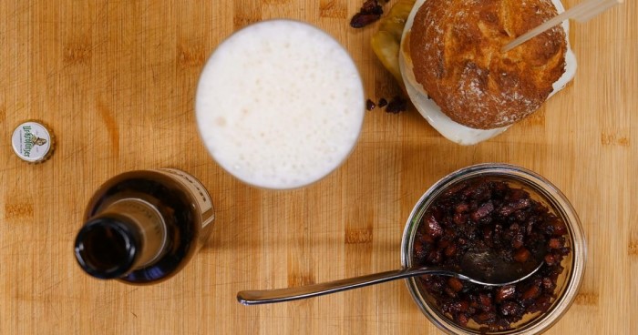 Ein Glas Bier steht neben einem Burger auf einer Holzoberfläche, daneben sieht man ein Glas mit Bacon.