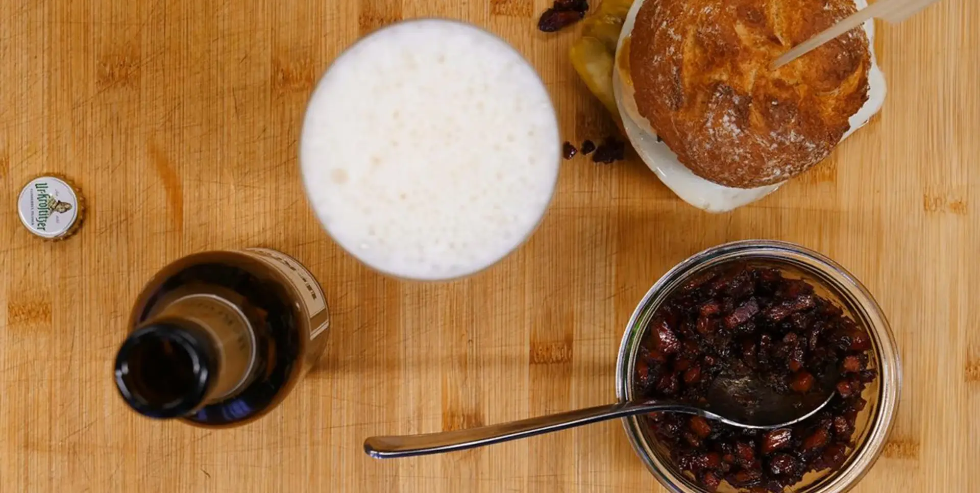 Ein Glas Bier steht neben einem Burger auf einer Holzoberfläche, daneben sieht man ein Glas mit Bacon.