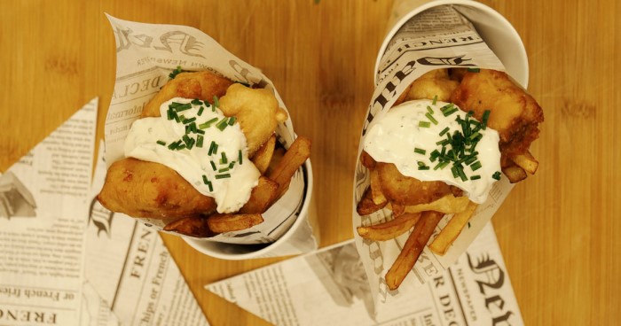 Zwei Portionen Fish and Chips in kegelförmigen Behältern stehen auf einem Holztisch.