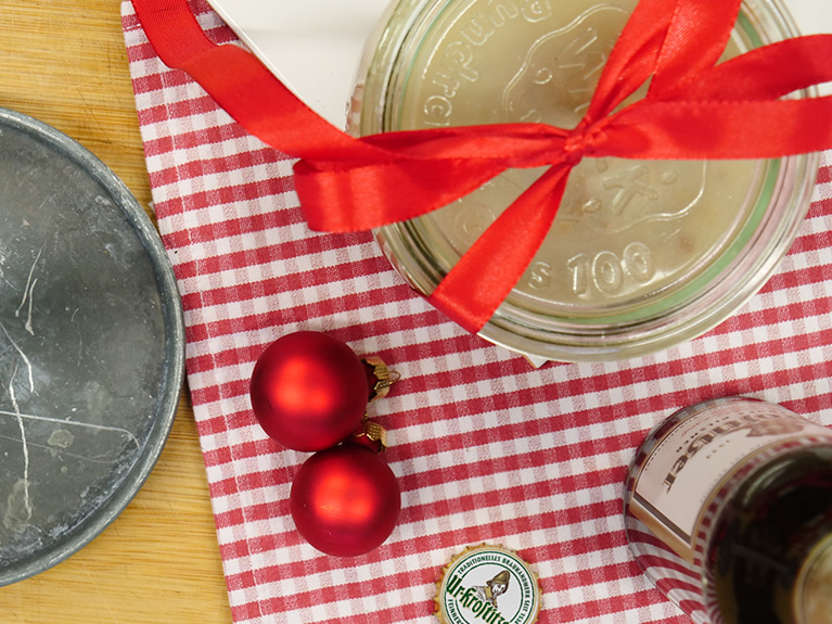 Zwei rote Weihnachtskugeln liegen neben einem Glas mit rotem Band auf kariertem Tuch.