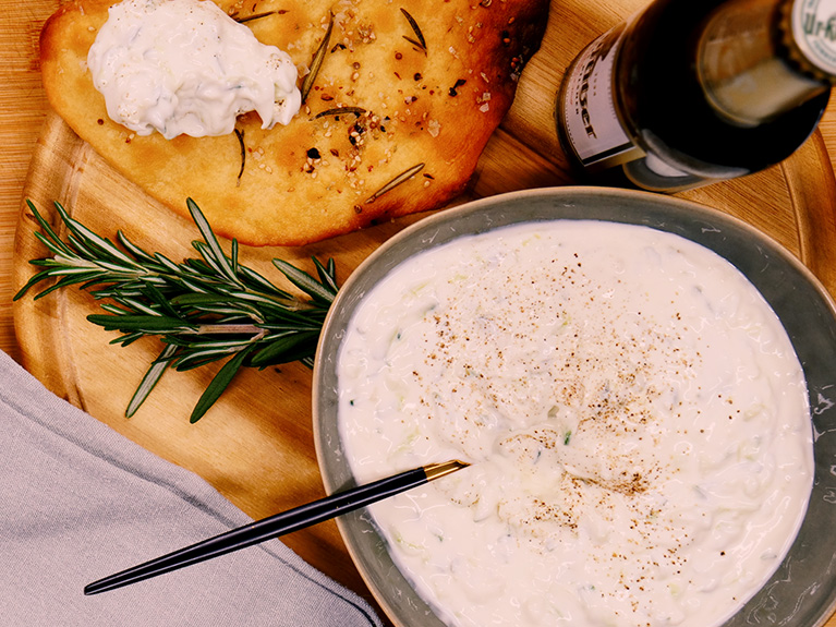 Eine Schüssel Tsatsiki steht neben einem Fladenbrot auf einem Holzbrett.