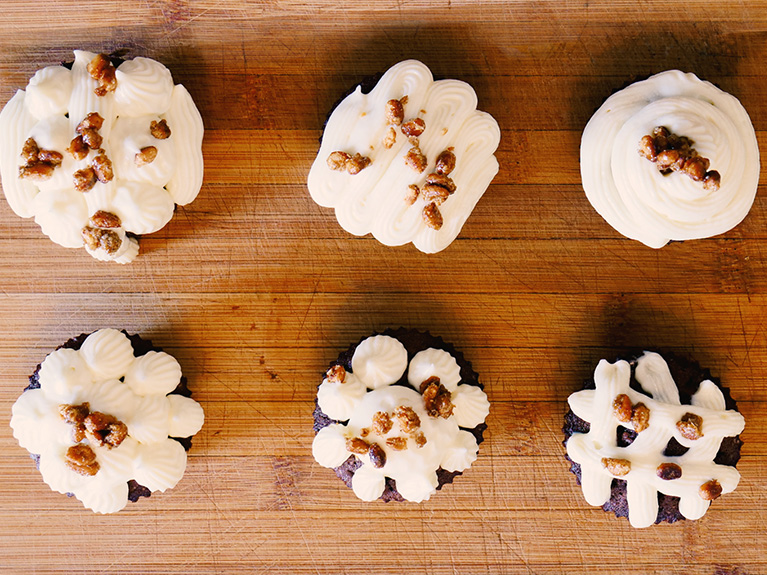 Sechs Schokoladenkuchen mit weißer Cremehaube und karamellisierten Nüssen sind auf einem Holzbrett zu sehen.