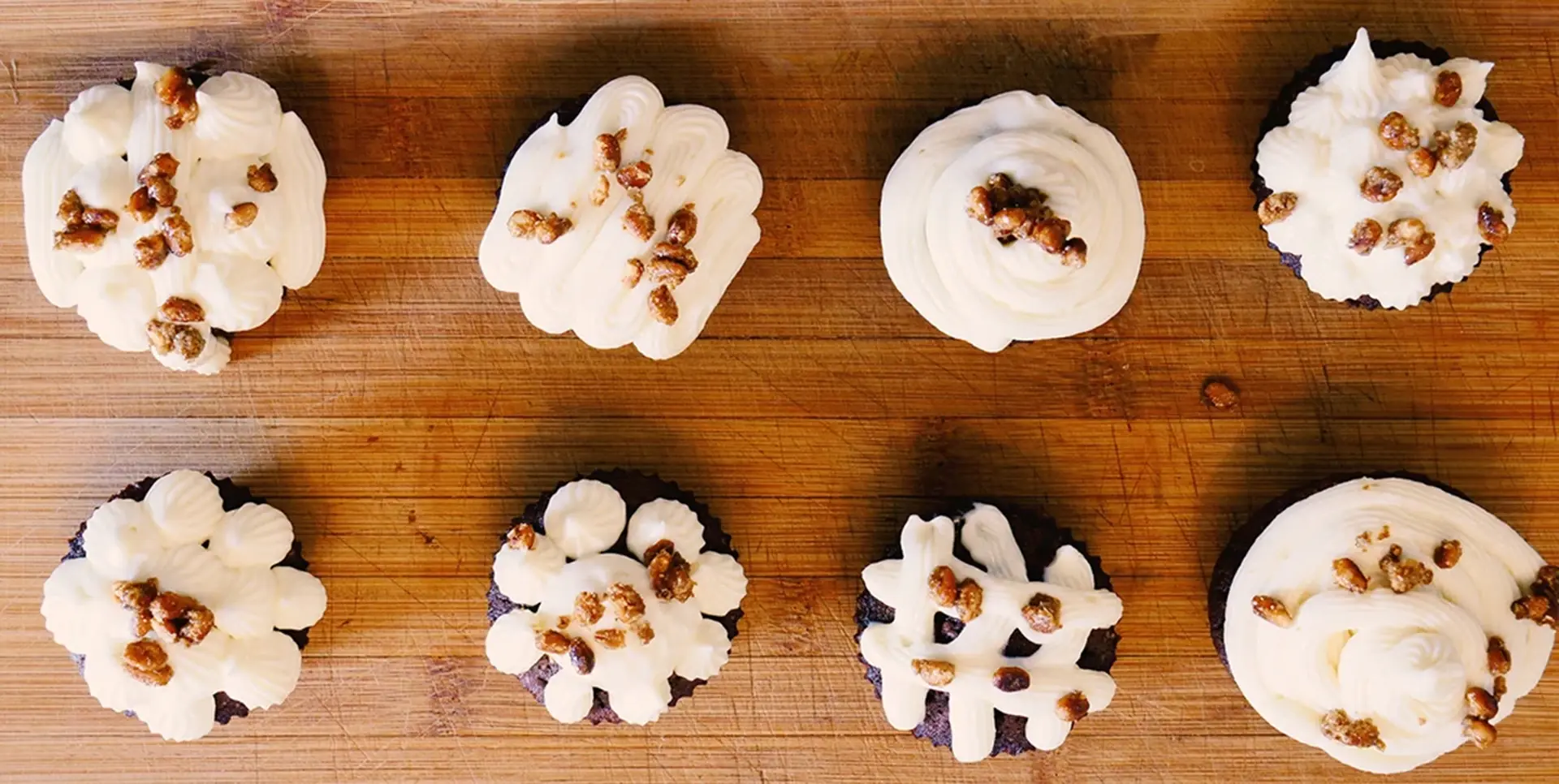 Acht Schokoladenmuffins mit weißer Creme und Nüssen sind auf Holzbrett angeordnet.