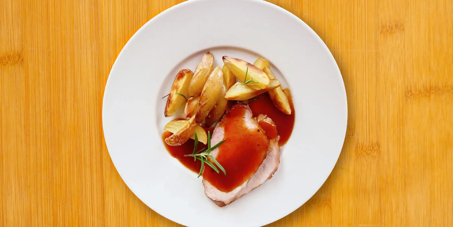 Ein Teller mit Schweinebraten und gebackenen Kartoffeln in Soße ist abgebildet.