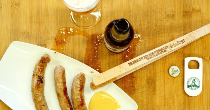 Drei Bratwürste liegen auf einem weißen Teller neben einem Glas Bier.