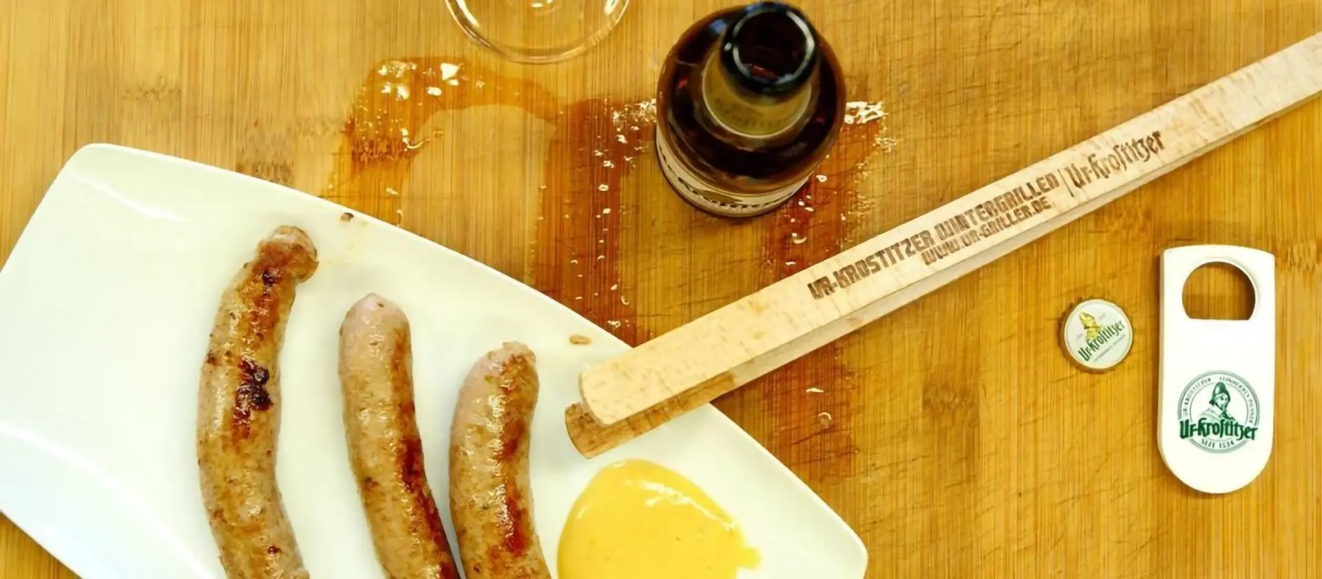 Drei Bratwürste liegen auf einem weißen Teller neben einem Bier. Ein Flaschenöffner und Bierdeckel sind auch zu sehen.