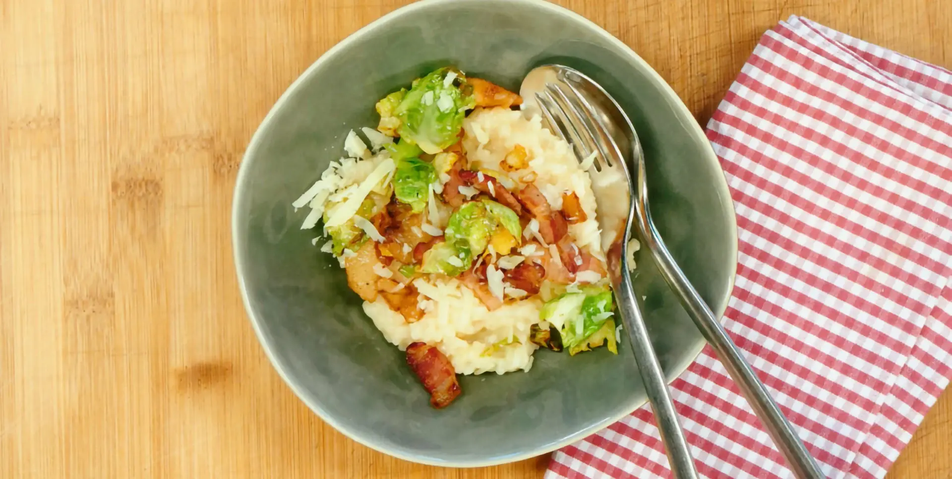Ein Teller mit Risotto, Rosenkohl und Speck, dazu eine Gabel auf einer Holzoberfläche.