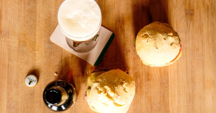 Ein Glas Bier steht neben zwei belegten Broten auf einem Holztisch.