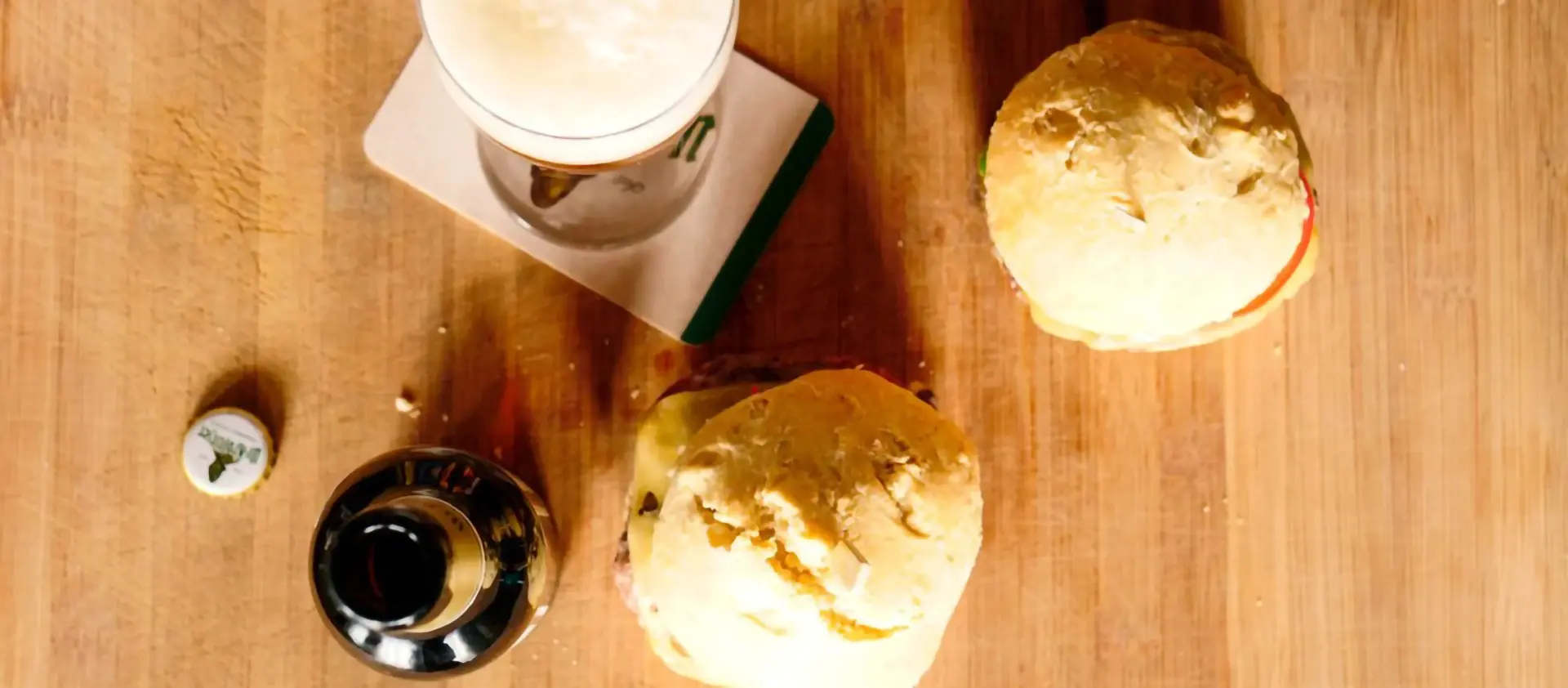 Ein Bierglas, eine Flasche und zwei belegte Brötchen sind auf einem Holztisch abgebildet.