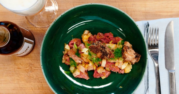 Ein grüner Teller mit Fenchelsalat und Rindfleisch steht auf einem Holztisch. Daneben liegen Besteck und ein Glas Bier.