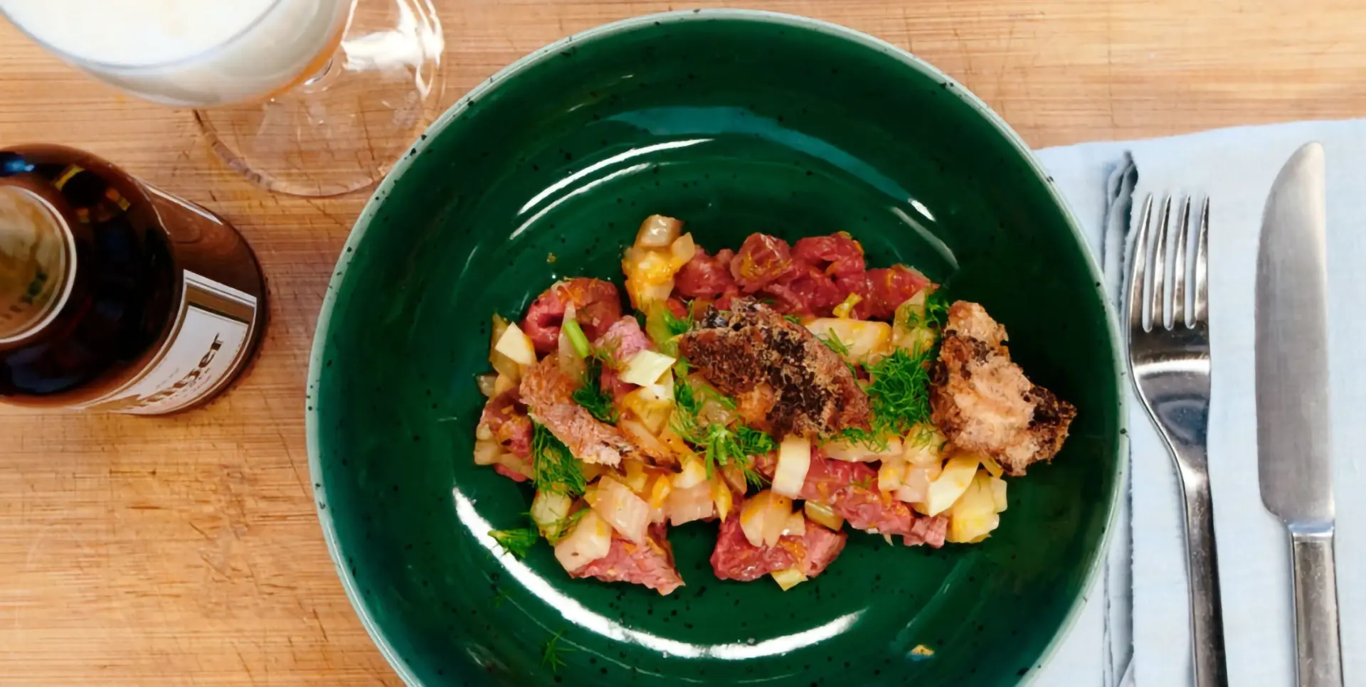 Ein grüner Teller mit Fenchelsalat und Rindfleisch steht auf einem Holztisch. Daneben liegen Besteck und ein Glas Bier.